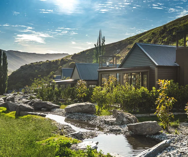 Des cabines avec des toits métalliques nichés dans un paysage de montagne luxuriant à côté d'un petit ruisseau par une journée ensoleillée.