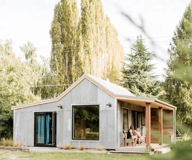 Tiny maison en métal ondulé avec un porche, entouré d'arbres, des gens assis sur le porche avec un chien à proximité.