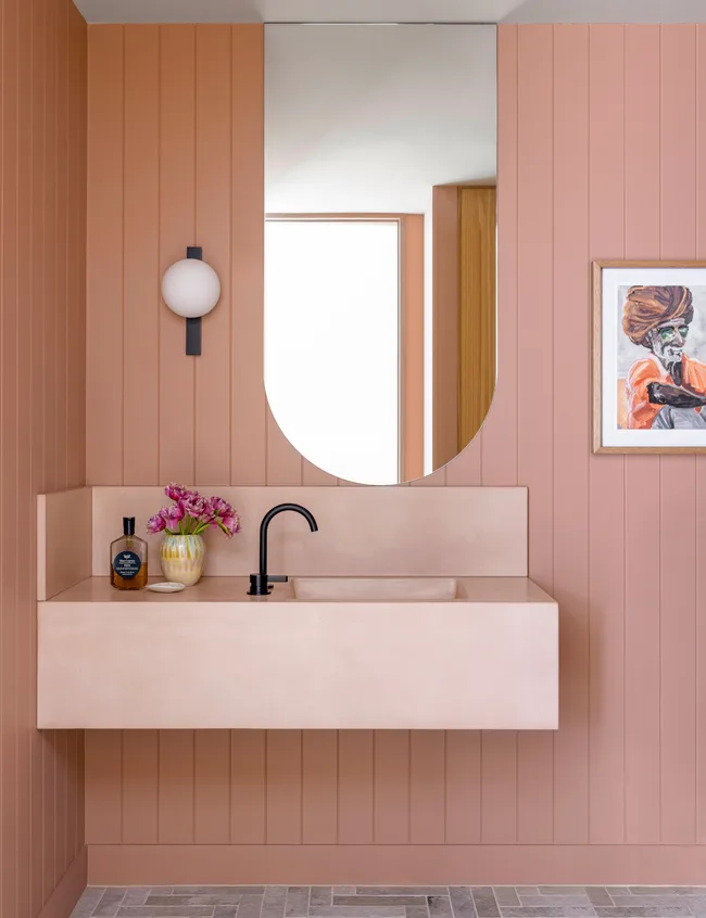 Salle de bain pêche avec lavabo mural, robinet noir, miroir rond, applexion murale, fleurs violettes et peinture encadrée.