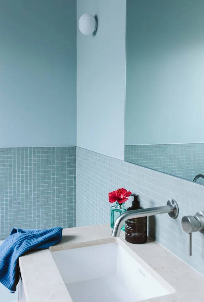Salle de bain moderne avec des murs bleus, des carreaux carrés, un robinet d'argent, une fleur rouge dans un vase et un distributeur de savon.