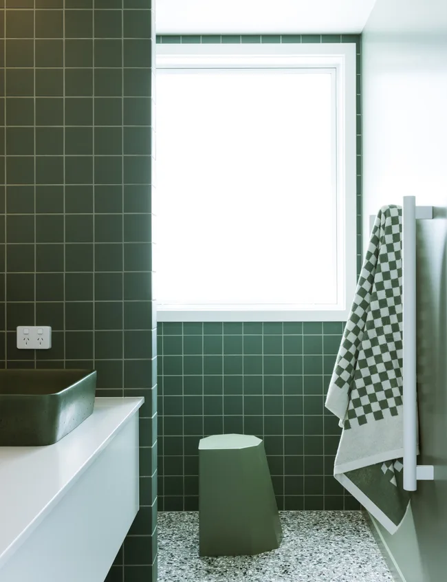 Salle de bain moderne avec carreaux verts, un lavabo blanc, tabouret géométrique et une serviette à carreaux sur une grille près de la fenêtre.