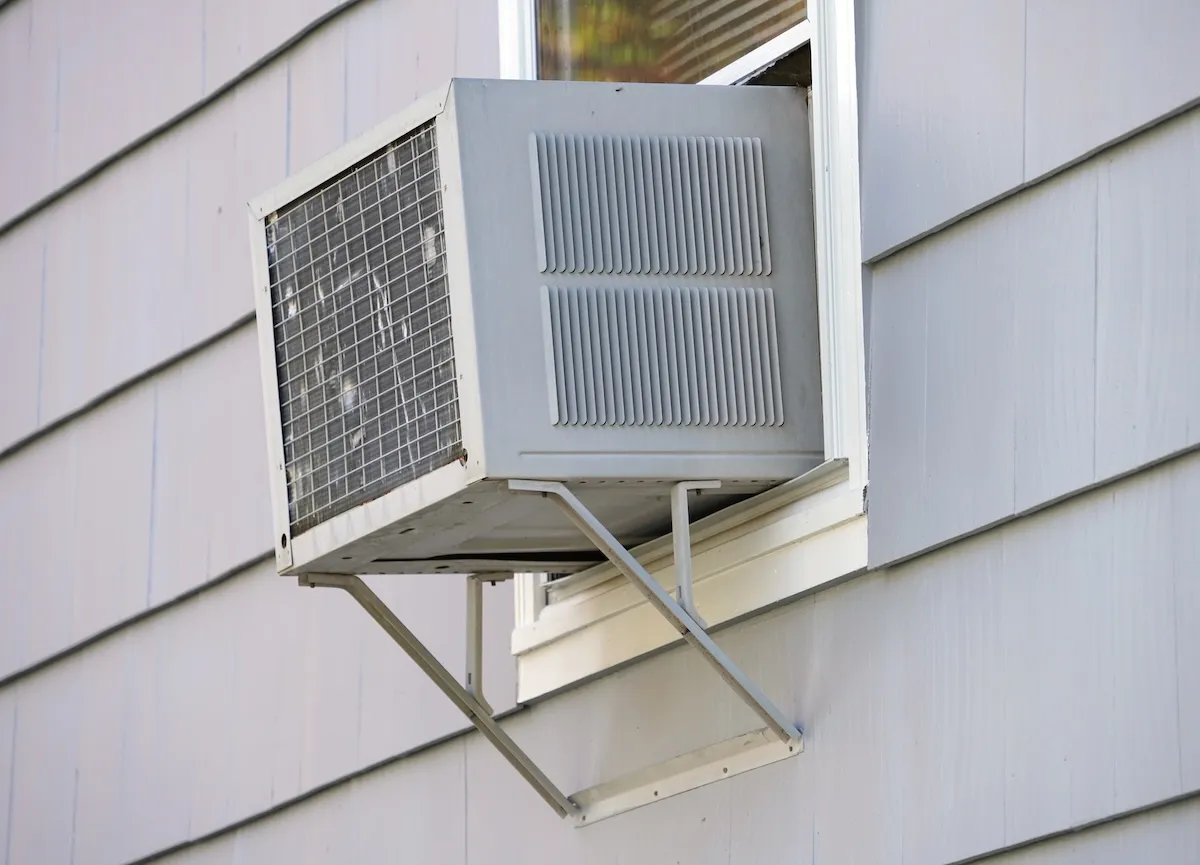 Old window air conditioner propped on a bracket on a home with grey siding.
