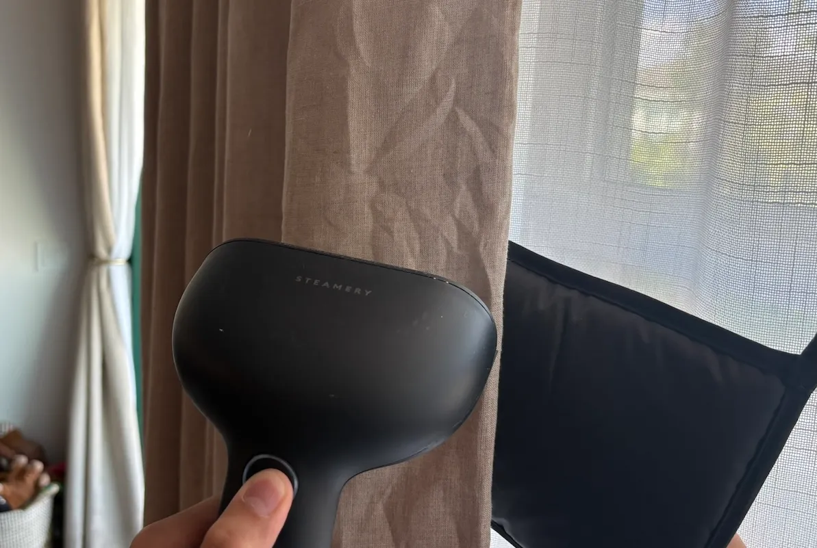 Person uses handheld steamer to steam wrinkles out of brown bedroom curtains.