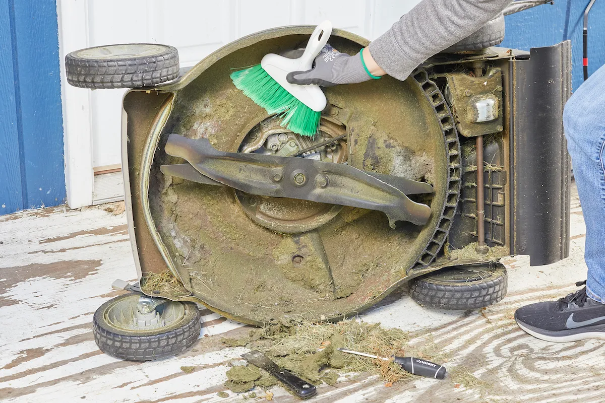 Person uses a hand broom to clean underside deck of lawn mower.