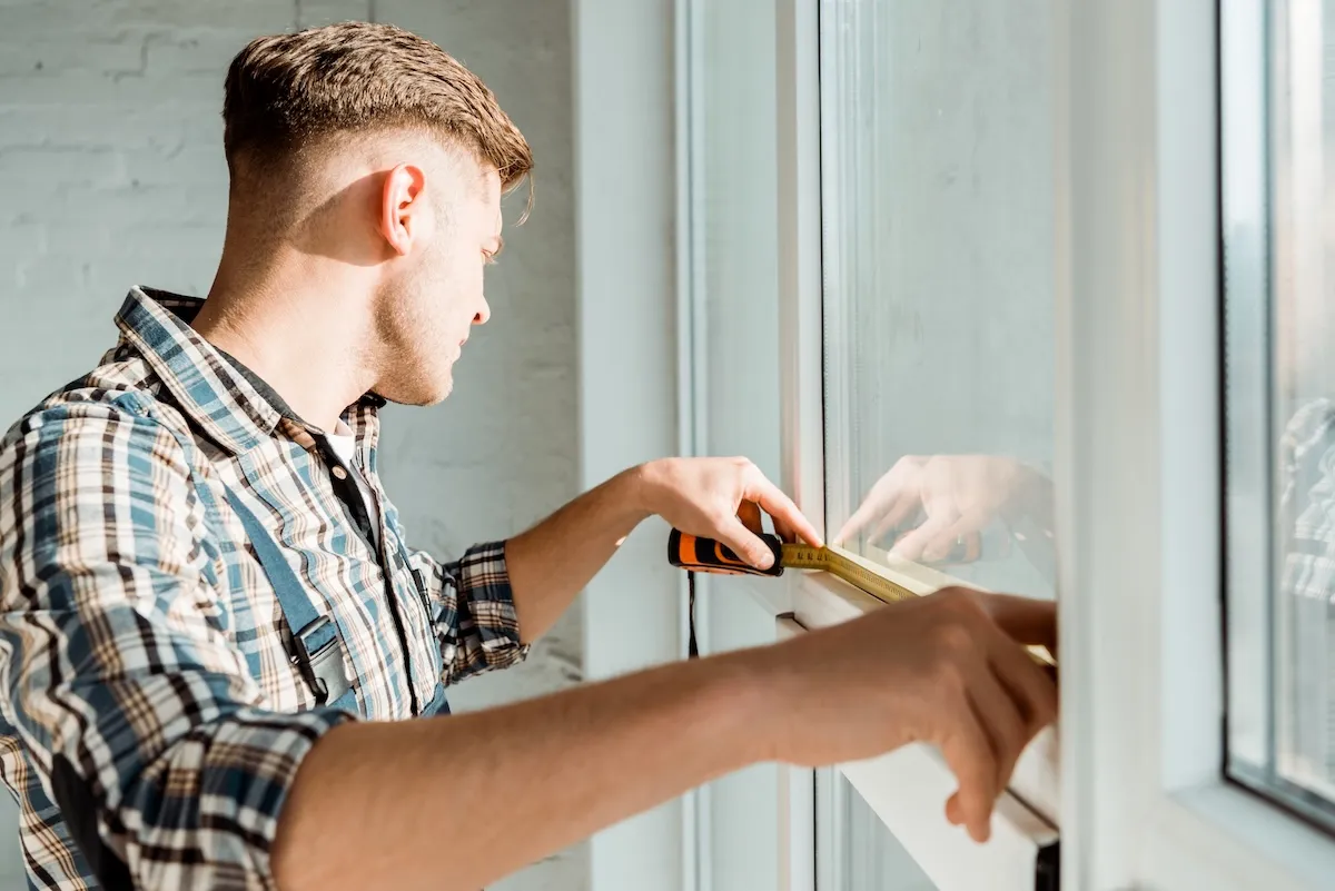 A man uses a tape measure to determine the dimensions of a sunny window. 
