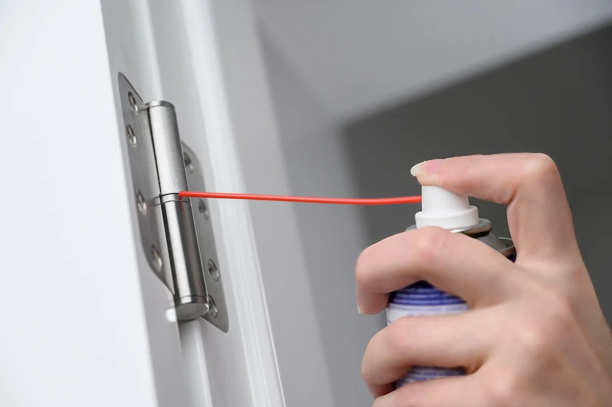 A man greases door hinges with a long straw pointed from the can at the hinges.