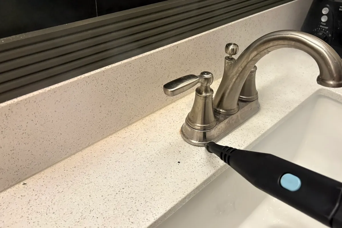 A steam cleaner's narrow nozzle cleans around the edges of a bathroom faucet.