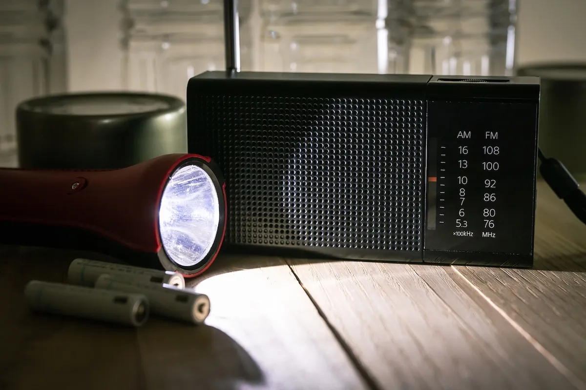 Flashlight lit, batteries, and radio on a table.