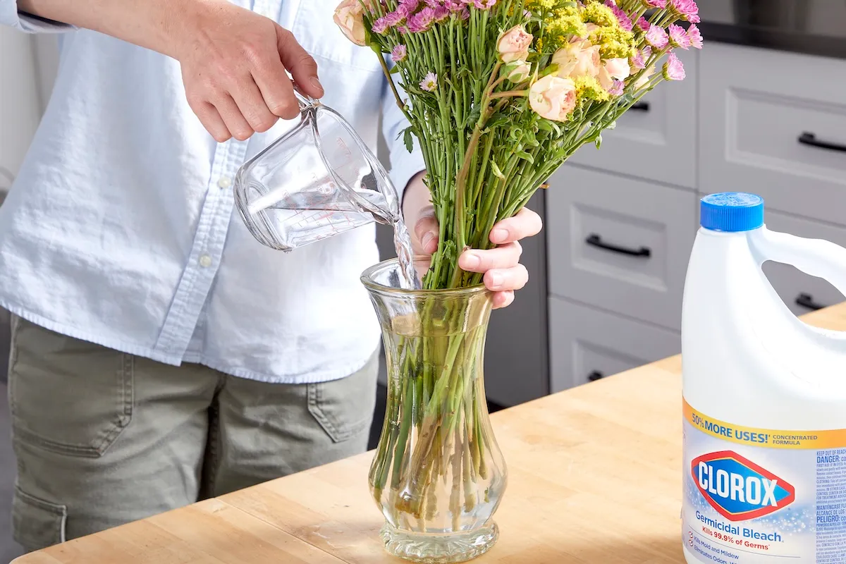 Woman pours water and bleach from a measuring cup in to a vase with cut flowers.