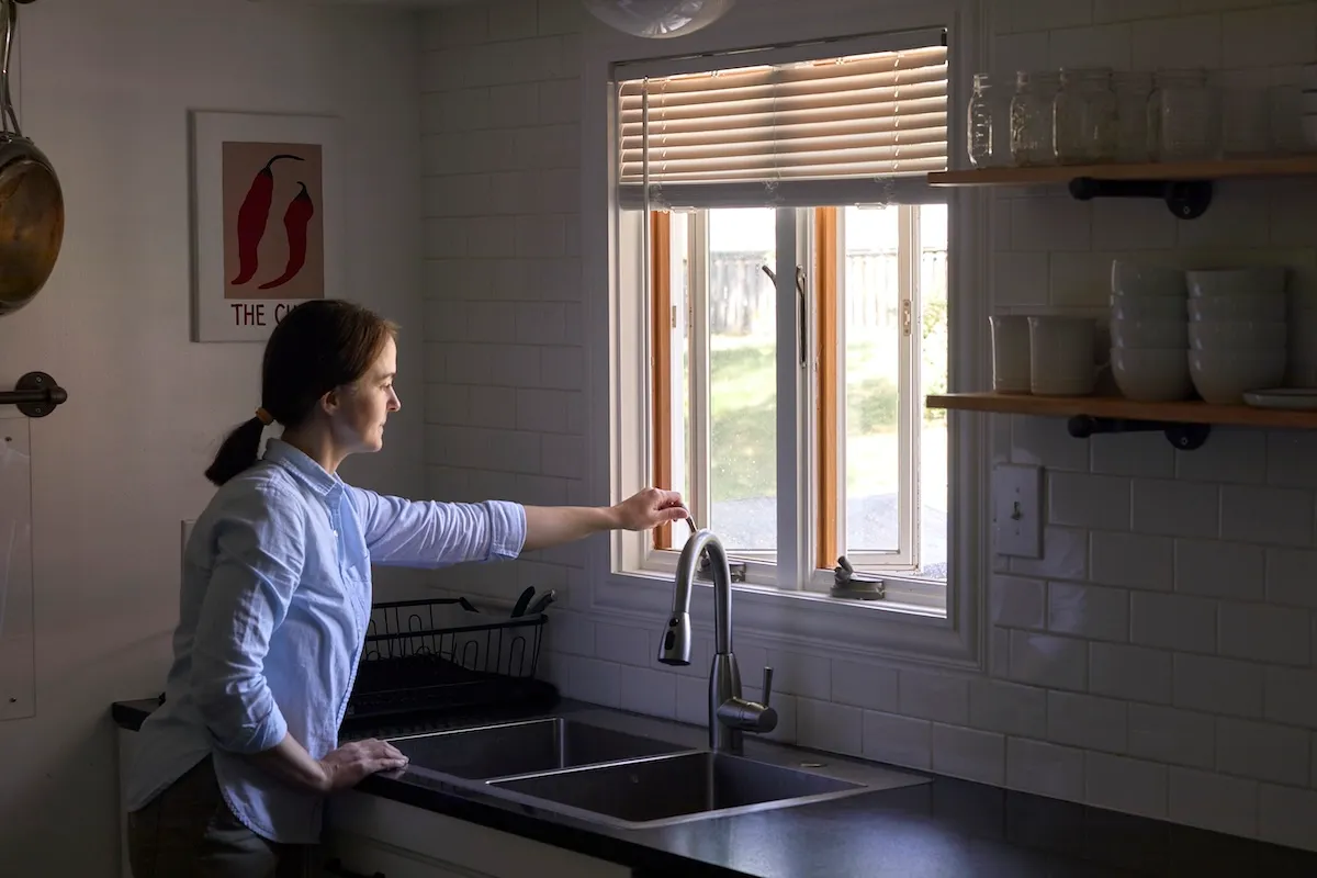 Woman in a dark kitchen cranks kitchen window open; window the only source of light.
