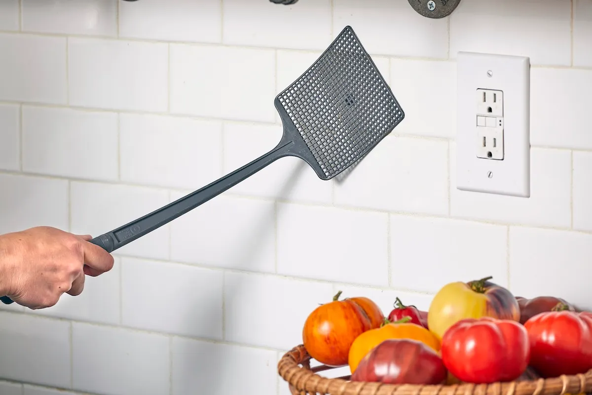Closeup of woman's hand holding a fly swatter against a kitchen backsplash to kill a fly.