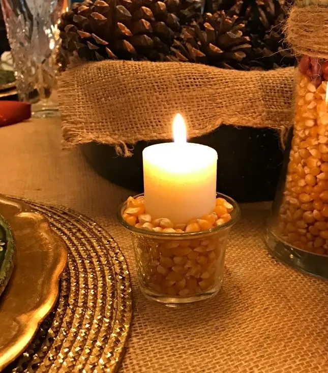 A lit votive candle arranged atop a jar of popcorn kernels lights an autumnal scene.
