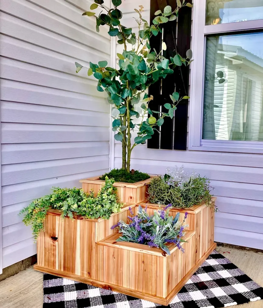 Front porch of house with white siding with a large square planter box with tiered sections with one small tree planted in the corner