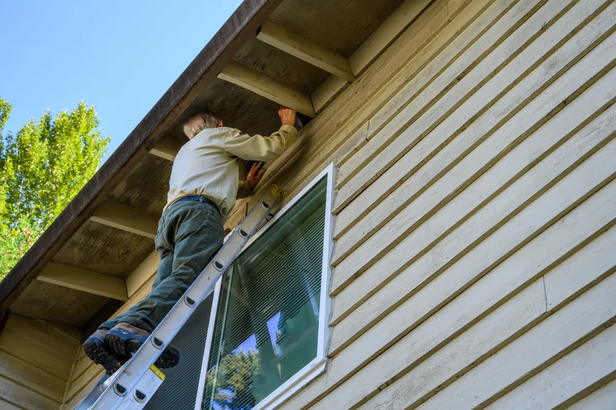 Man on a ladder checks attic vents under roof eaves.