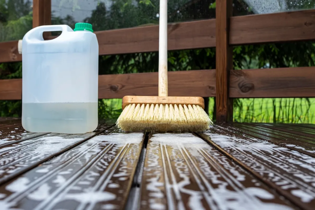 A deck scrub brush and a plastic container of cleaning solution sit on a soapy wood deck.