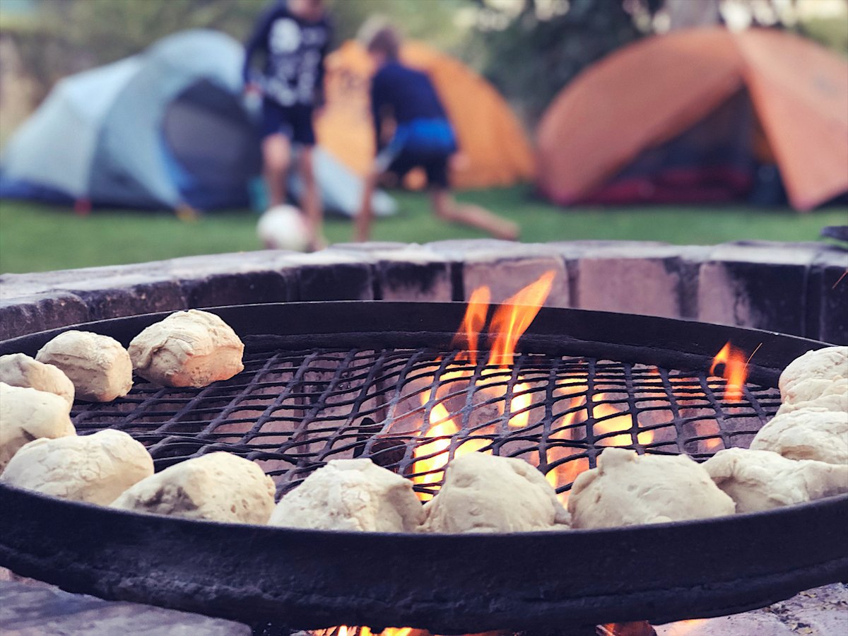 Metal fire pit with a grate upon which food is being cooked over a roaring fire.