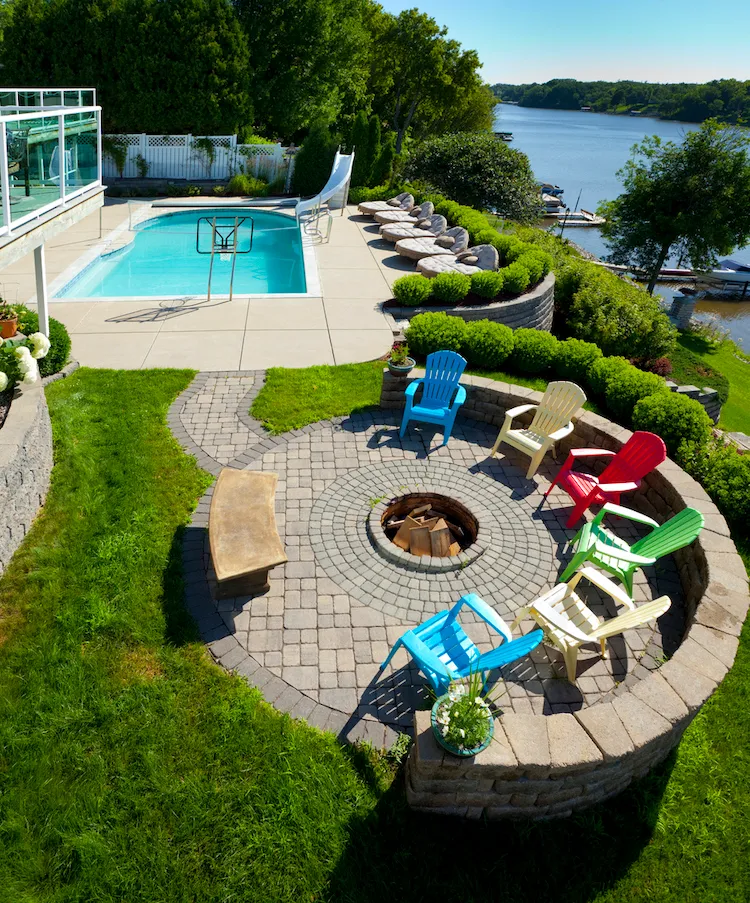 High view looking down on a built-in firepit surrounded by colorful Adirondack chairs; a swimming pool in the background.