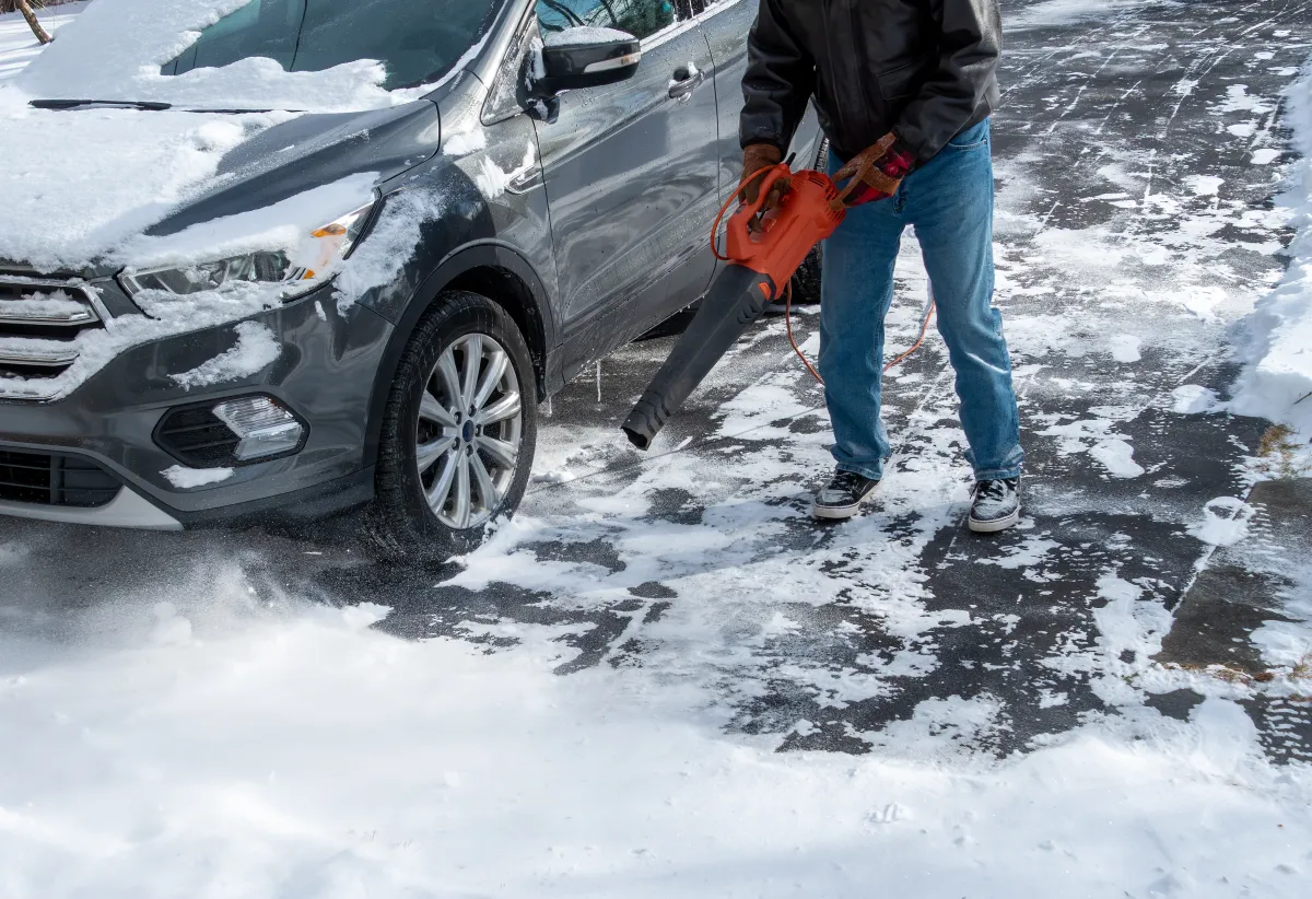 A man using a leaf blower to clear light powdery snow from car and the driveway