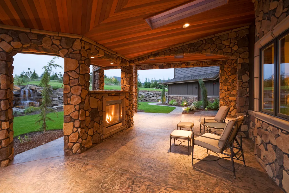 A covered backyard patio features a stone fireplace fueled by stone, chairs, and small tables.