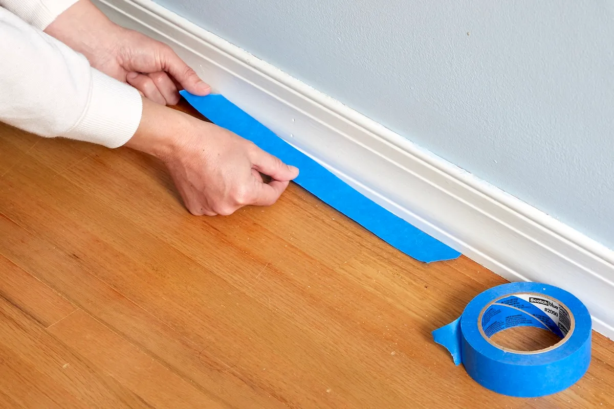 Woman applies painter's tape next to baseboard to protect hardwood floor.