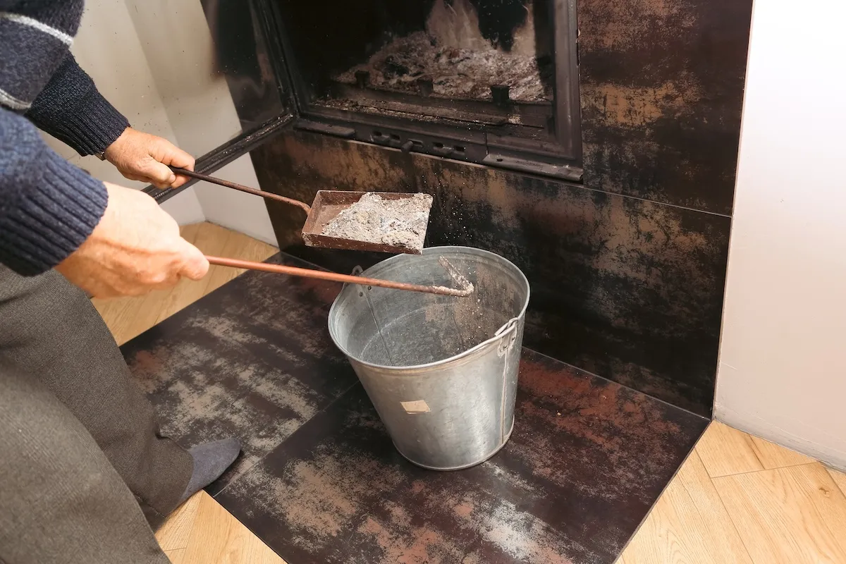 A man uses a small scoop and ash bucket to clean his residential fireplace.