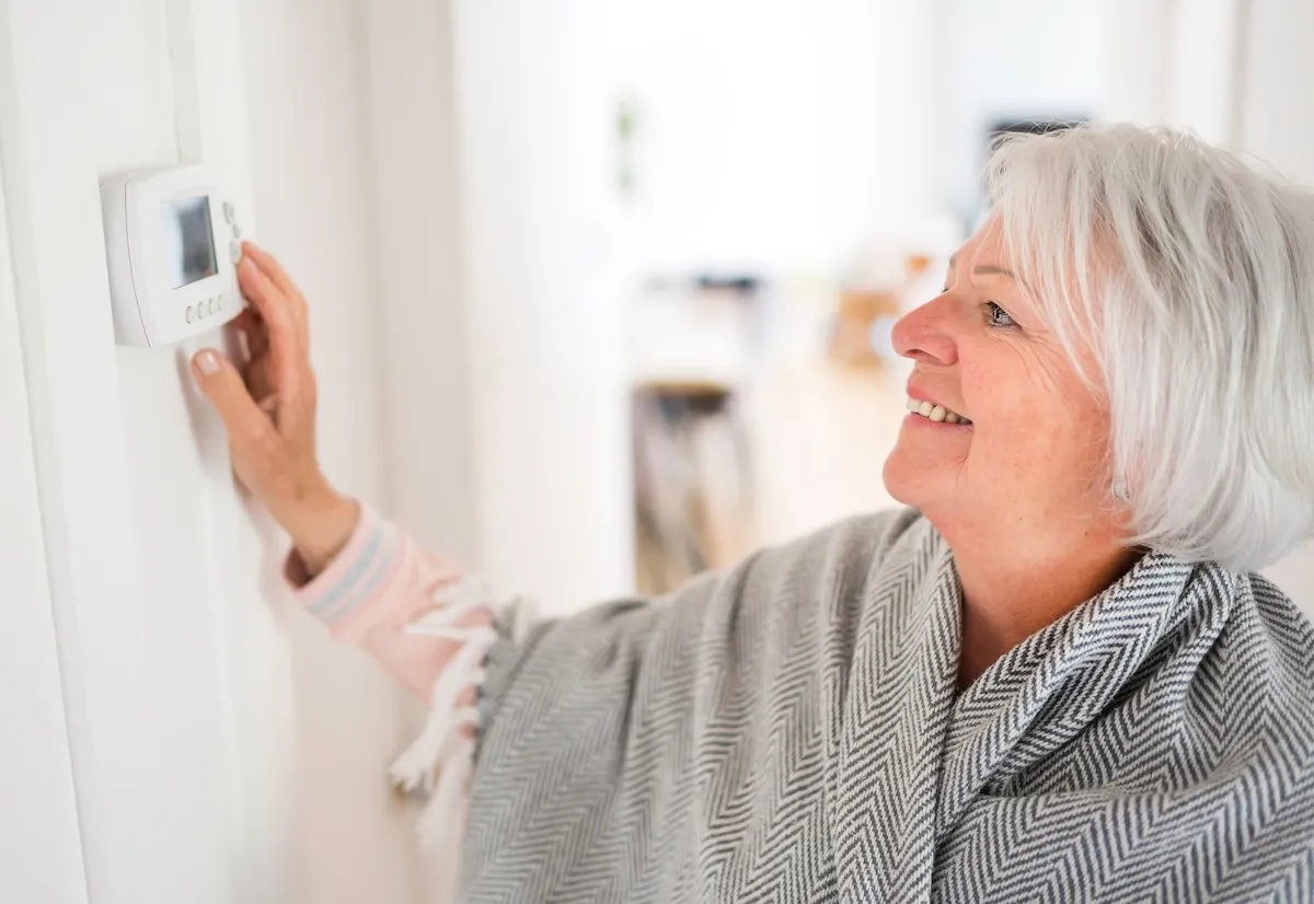 A mature woman wrapped in a blanket adjusts her thermostat. 