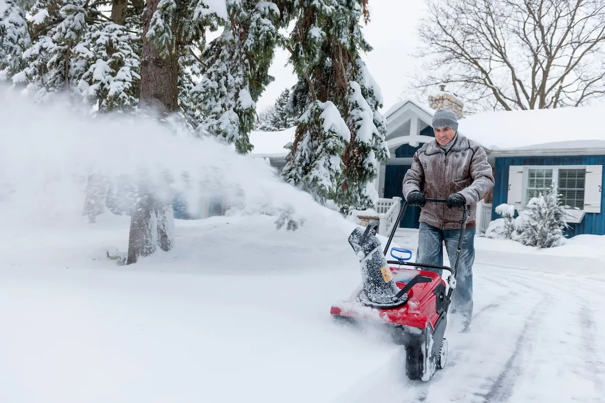 A man wearing snow gear uses a snow blower to clear his snowy driveway.