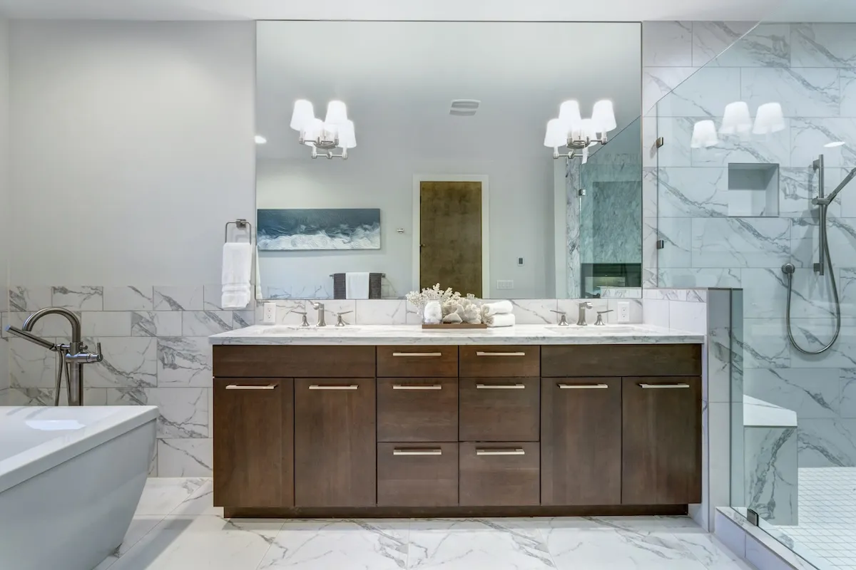 Luxurious master bathroom with Carrara marble tile surround, modern glass walk in shower, espresso dual vanity cabinet with a large mirror, and a freestanding bathtub.