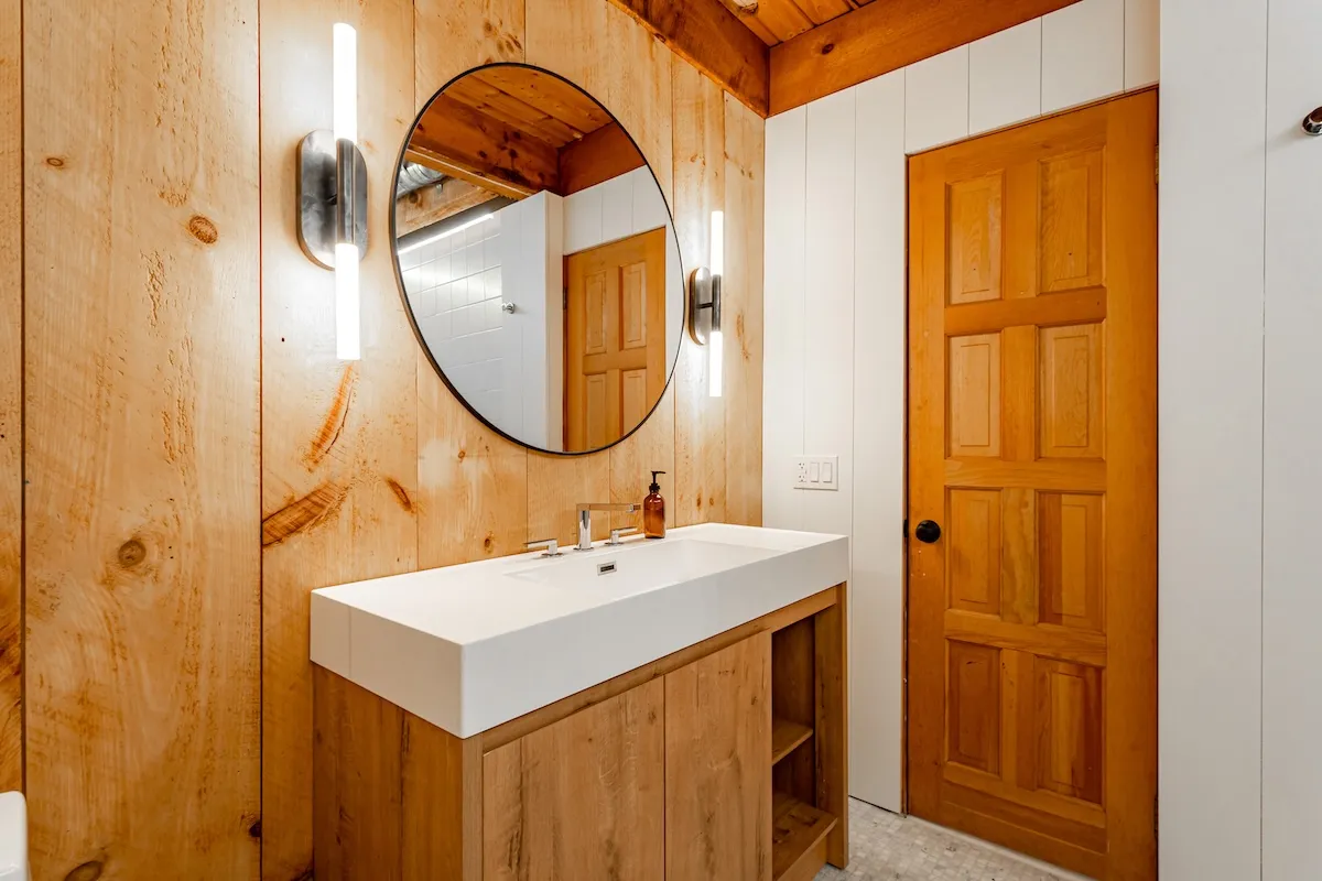 Rustic, cozy, wood-paneled bathroom with a round mirror, single vanity, and sconces next to the mirror.