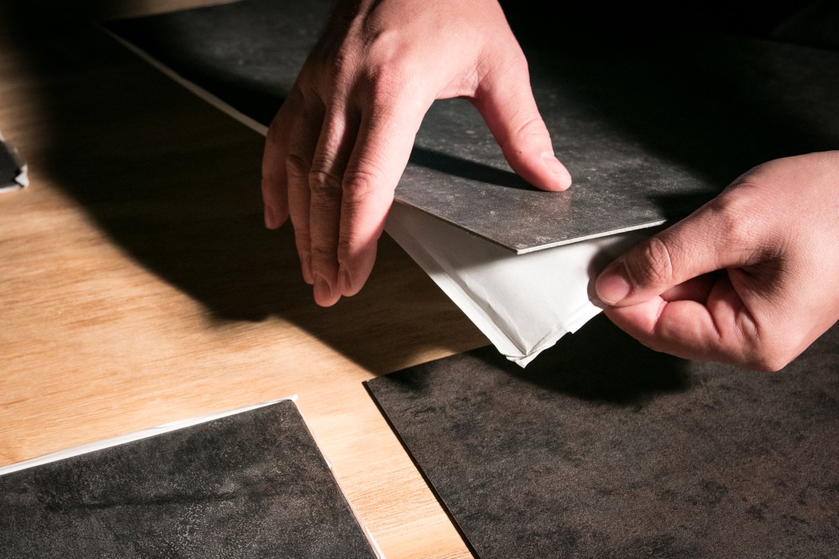Person peels the plastic backing off dark grey adhesive vinyl flooring tiles.
