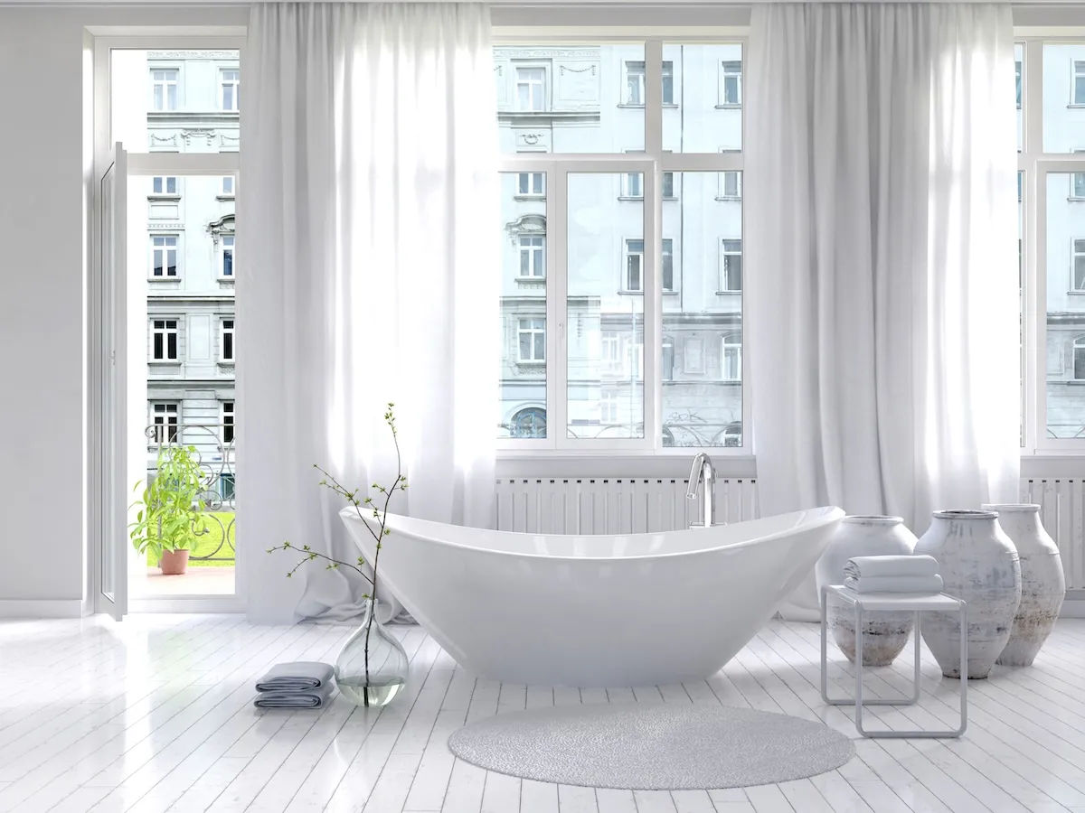High-ceilinged all-white bathroom with a wall of windows, sheer white curtains, and a freestanding white bathtub.