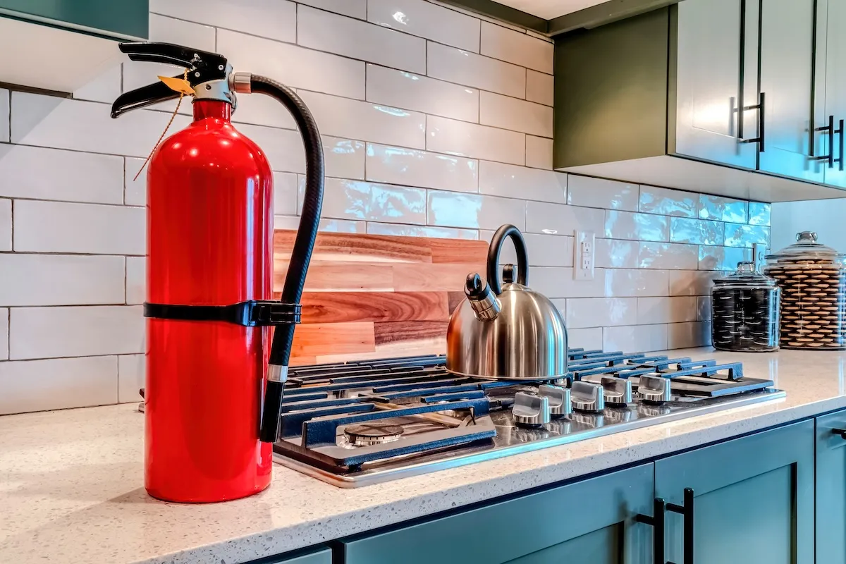 Fire extinguisher and cooktop with kettle on the countertop inside home kitchen.