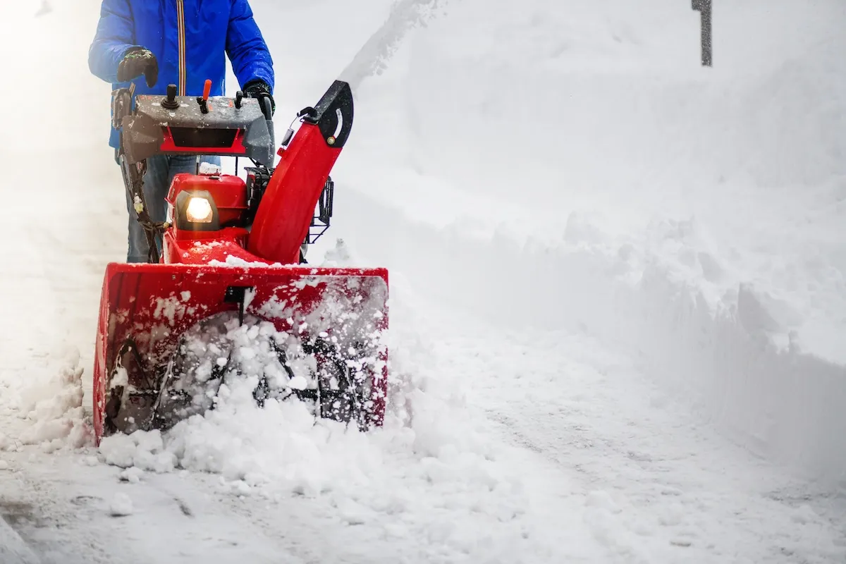 A man in a blue jacket clears snow from a driveway with a large snowblower.
