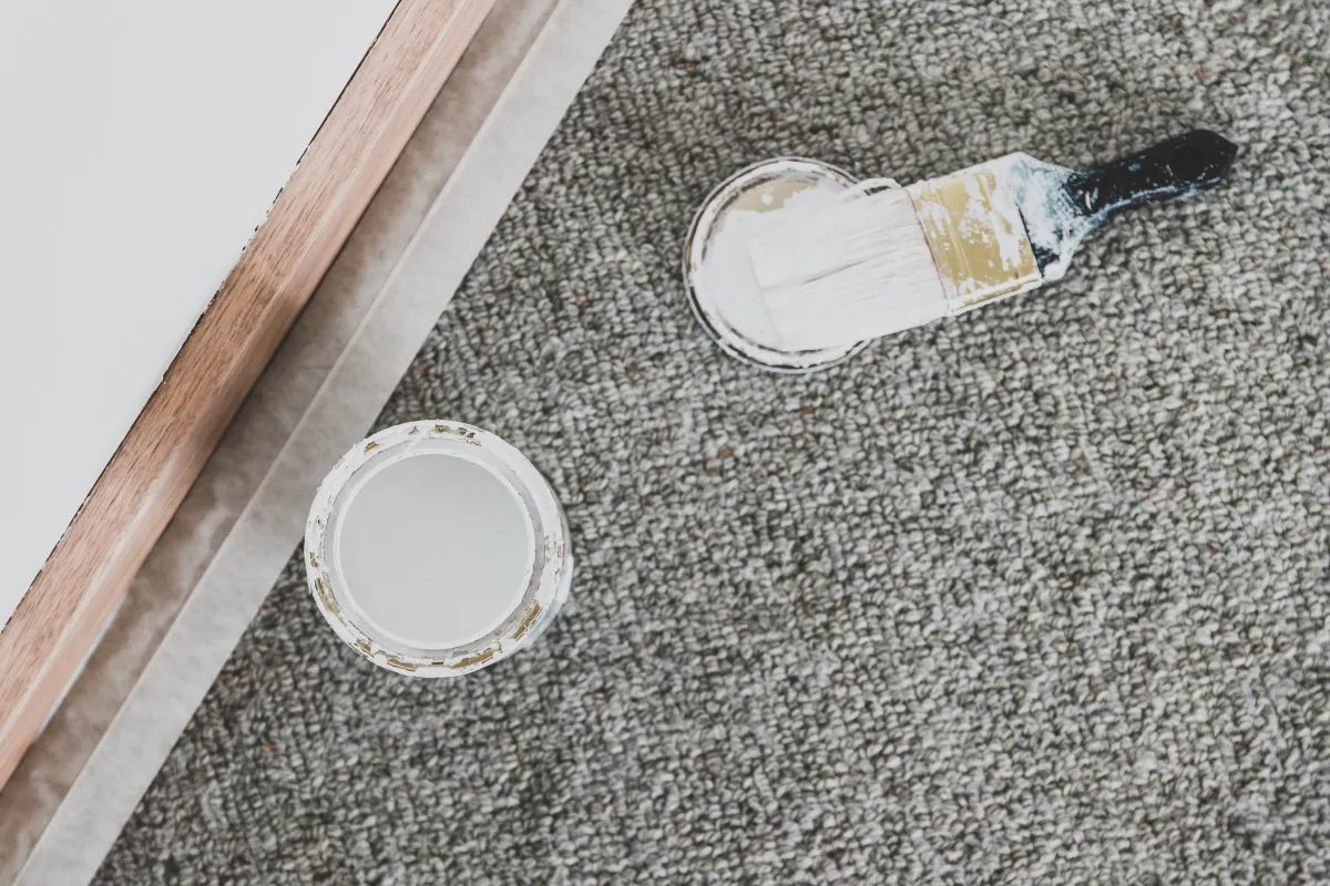 Open can of white paint on a carpet with a paintbrush resting on the lid.