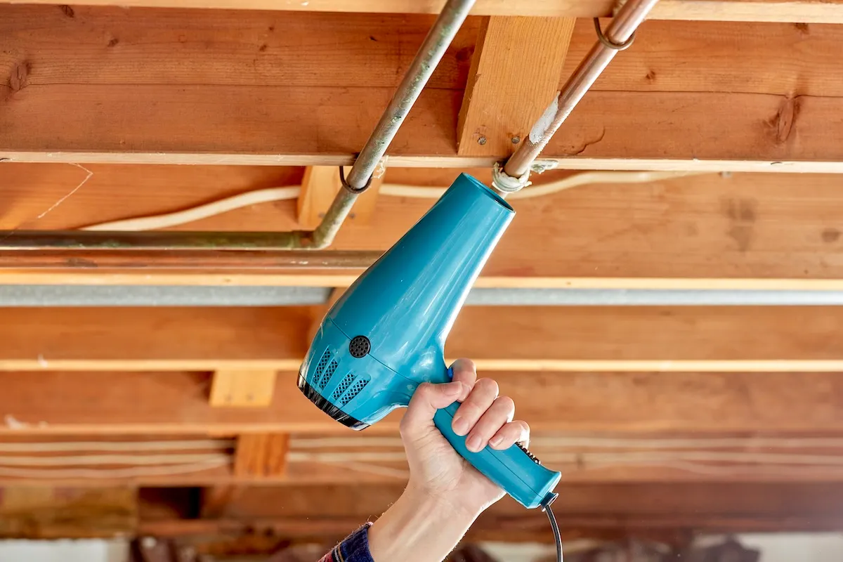 A DIYer using a hair dryer to thaw a frozen pipe in an unfinished basement.